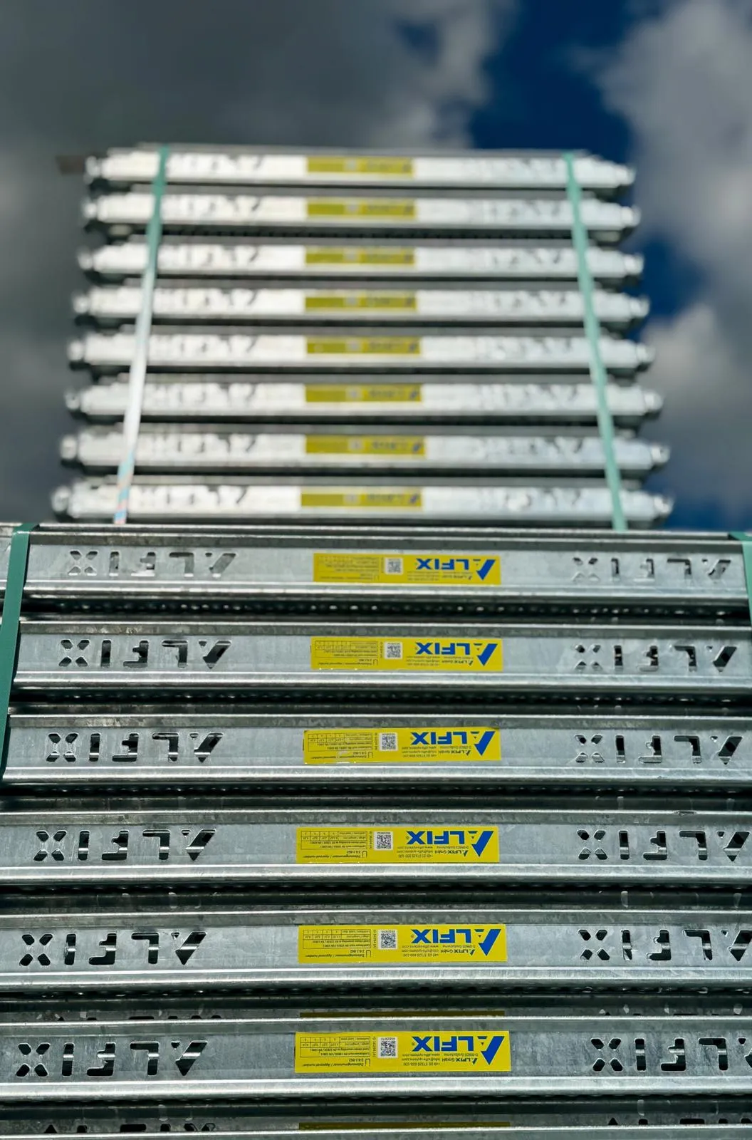 Metallisten ALFIX-rakennustelineiden pino, sidottu vihreällä nauhalla, taustalla sininen taivas ja pilvet.