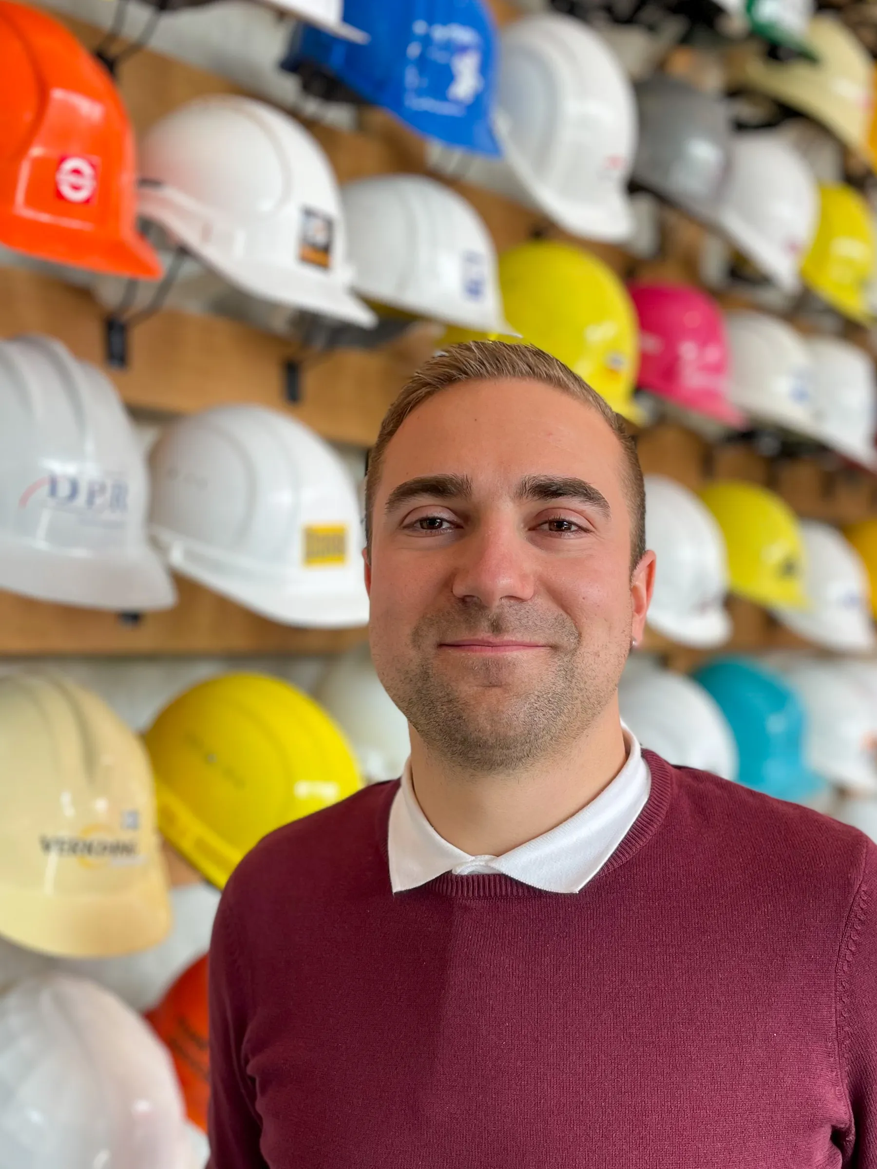 Mies seisoo seinän edessä, jossa on rivi värikkäitä suojakypäriä. Hänellä on yllään viininpunainen villapaita ja valkoinen kauluspaita. Valo on kirkas ja tasainen.