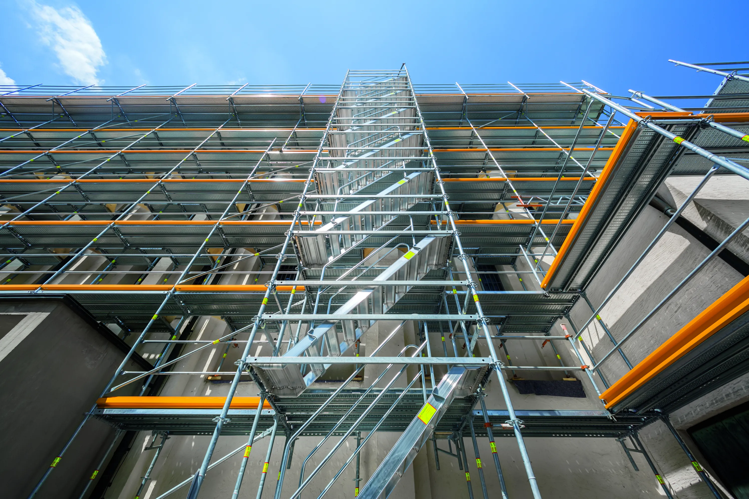 Korkea rakennusteline sinistä taivasta vasten, metallinen rakenne oransseilla suojakaiteilla ja harmaalla rakennuksella taustalla, kirkas valaistus.