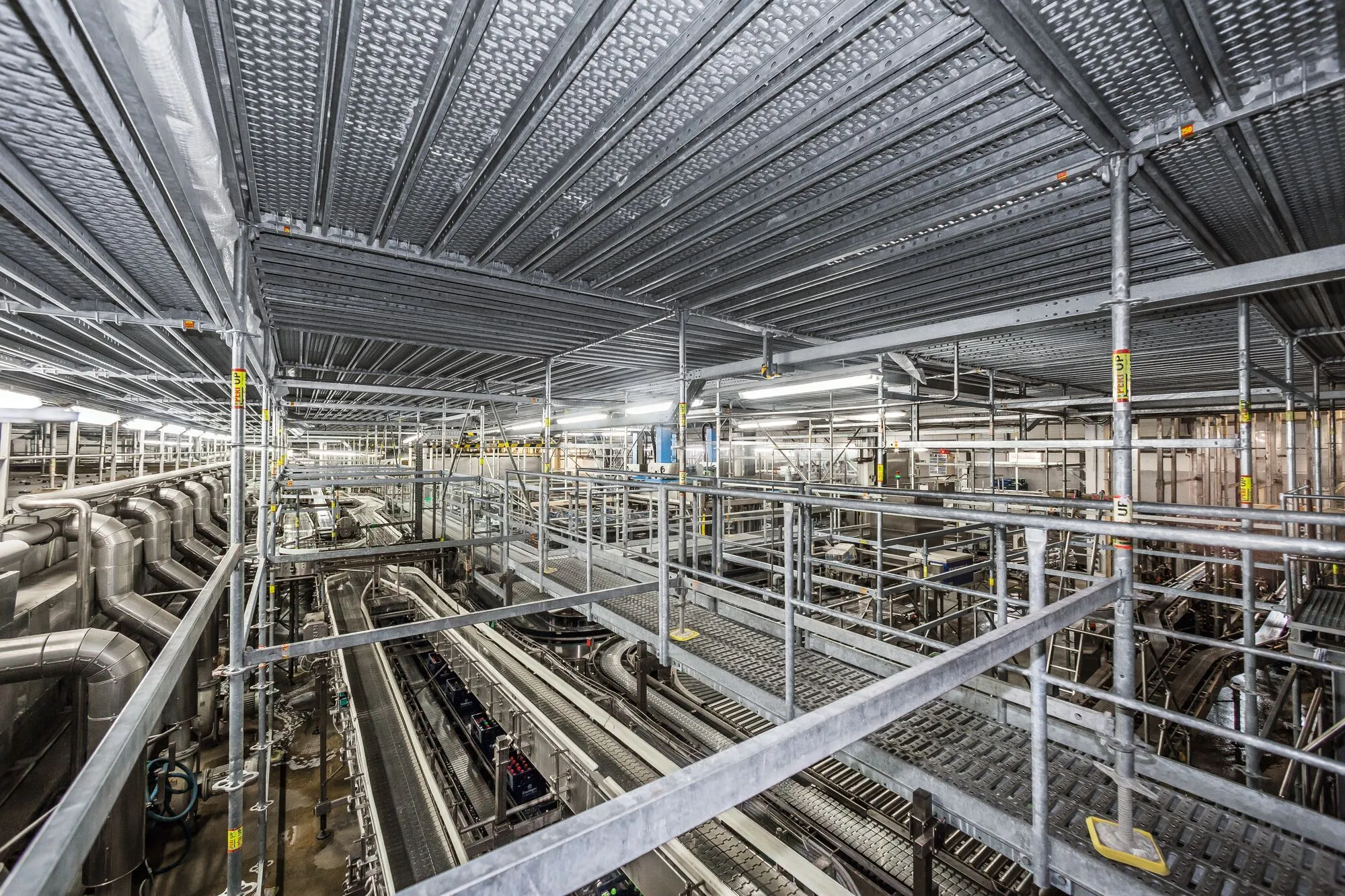 Teollinen sisätila, jossa on metalliputkia, kuljettimia ja valaistus, luoden monimutkaisen ja teknisen näkymän.