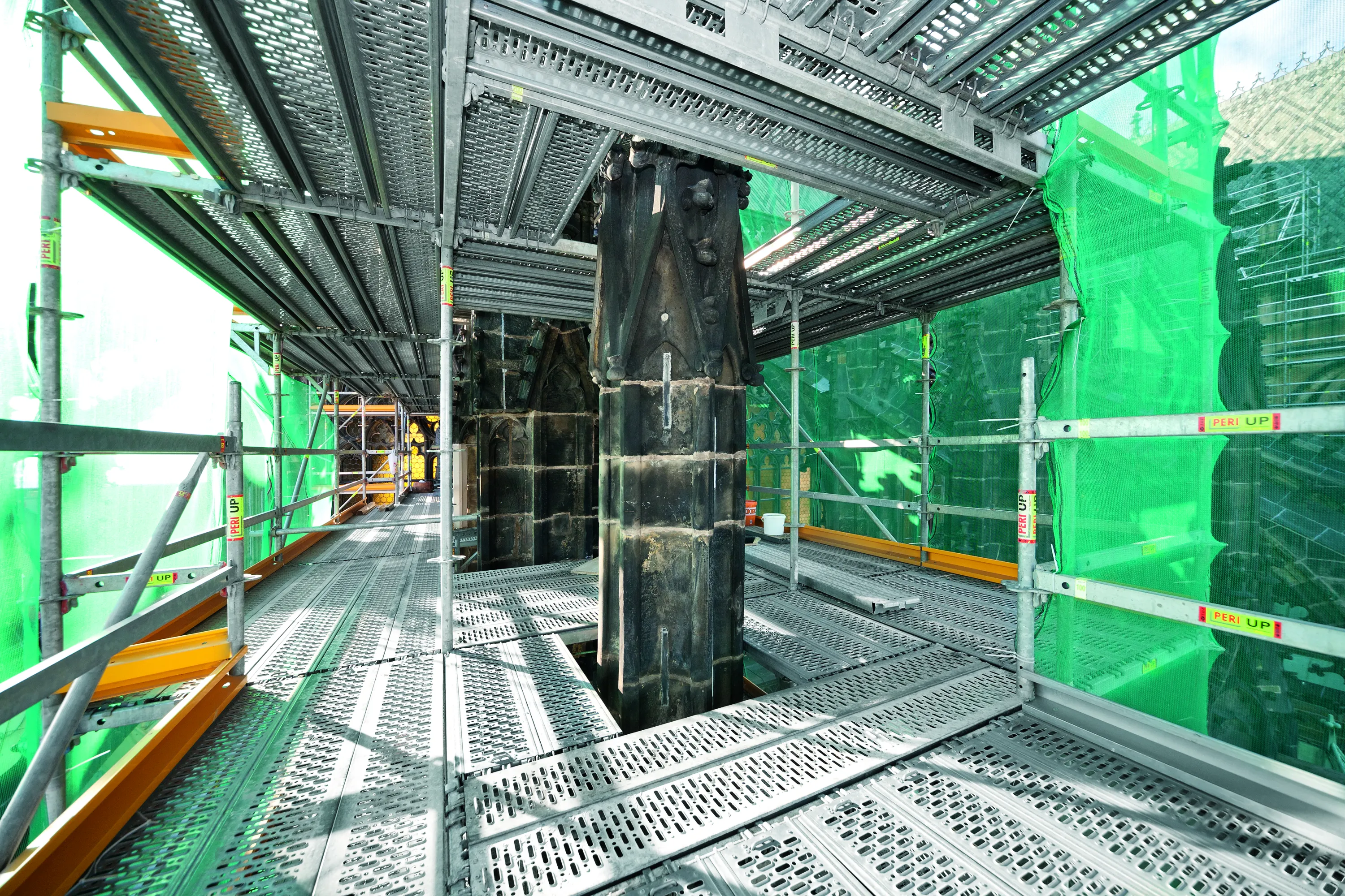 Rakennustelineet ympäröivät tummia kivipilareita, vihreä verkko peittää osan telineistä, valoisa ja metallinen tunnelma.